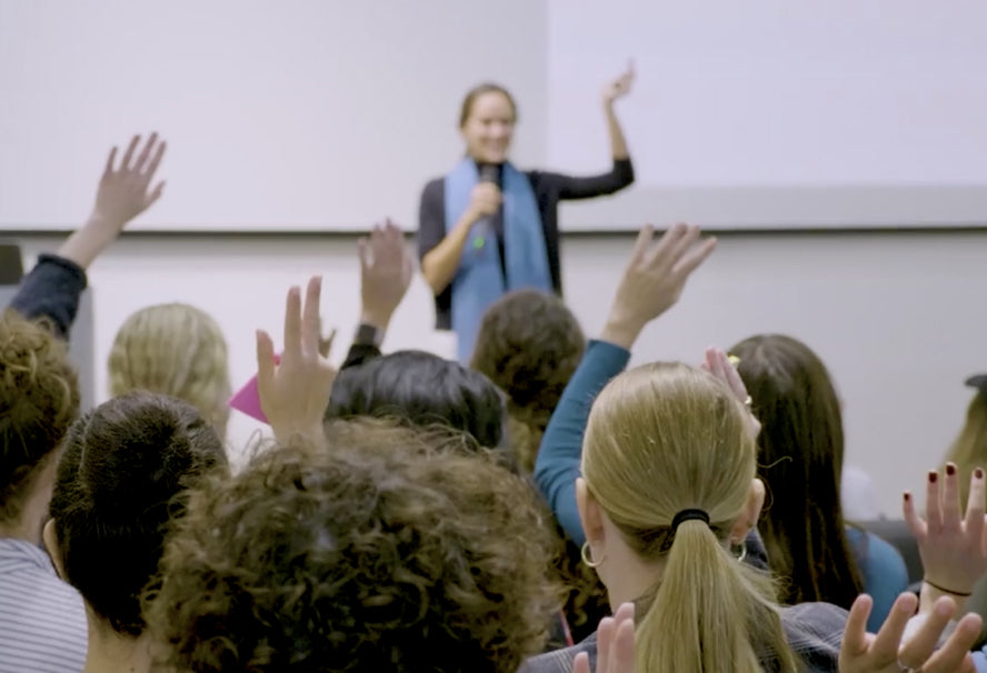 A woman standing in front of a group giving a presentation with one hand raised and many people in the audience have one hand raised. 